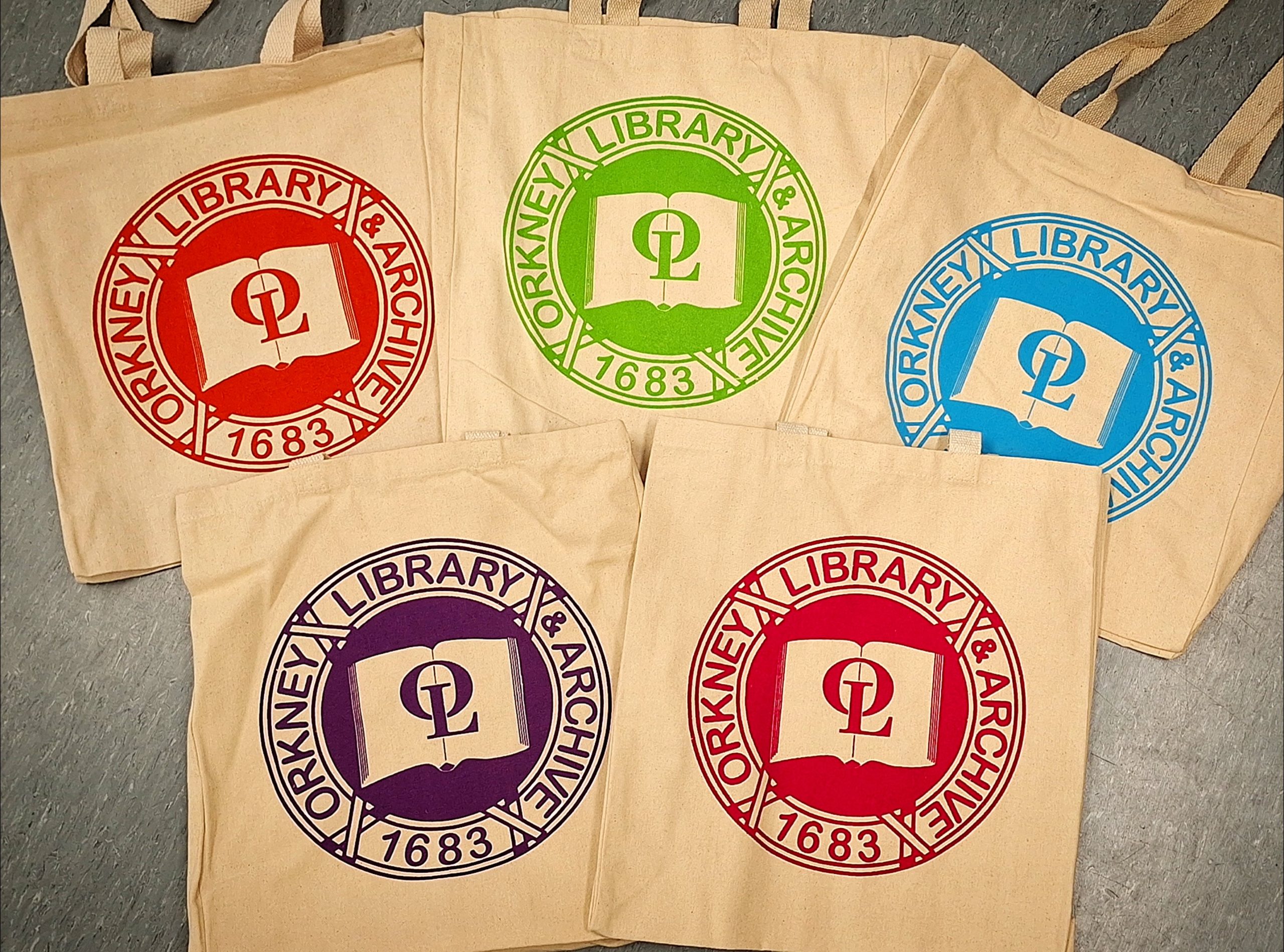 Five tote bags displayed with the Orkney Library & Archive logo in red, green, blue, purple or pink