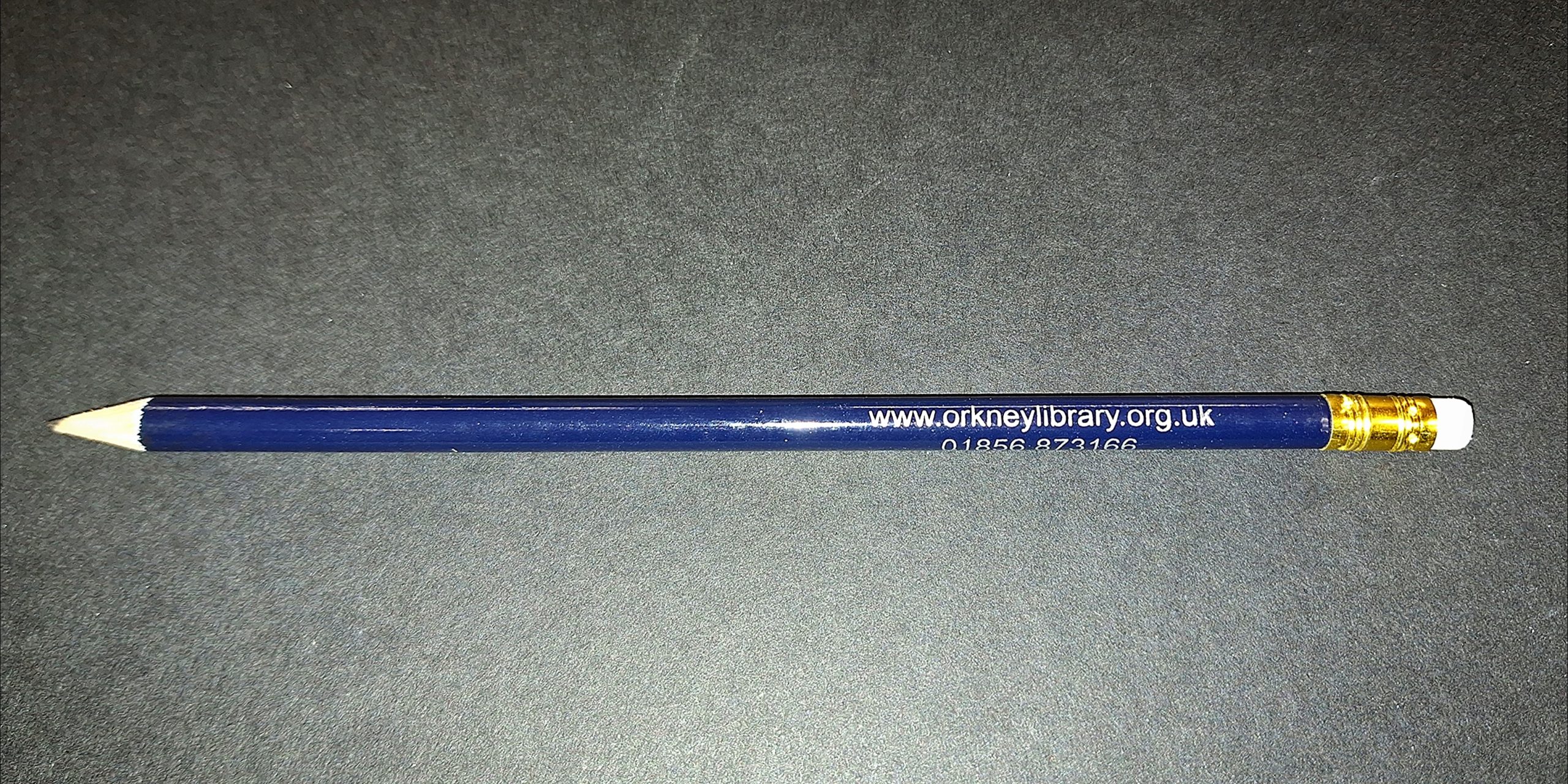 A blue pencil with a white eraser and gold ferrule, featuring the text "www.orkneylibrary.org.uk 01856 873166" printed in white along the side, placed against a dark gray background