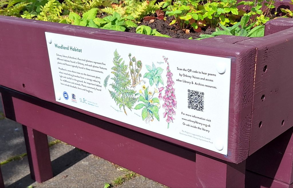 A wooden planter filled with flowers. There is an information panel with a QR code.